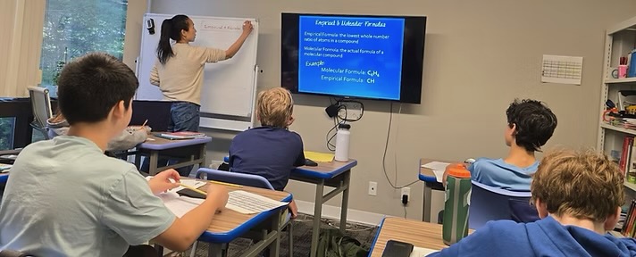 A WSA teacher leading a chemistry lesson on empirical and molecular formulas while students take notes at their desks